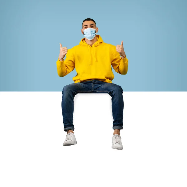 Informace o očkování. Šťastný arabský muž v ochranné lékařské masce sedí na prázdném štítku a ukazuje palce nahoru — Stock fotografie