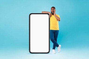 Şok olmuş siyah bir adam, büyük akıllı telefonun yanında duruyor. Uygulamanız ya da web siteniz için mavi stüdyonun arka planında bir model.