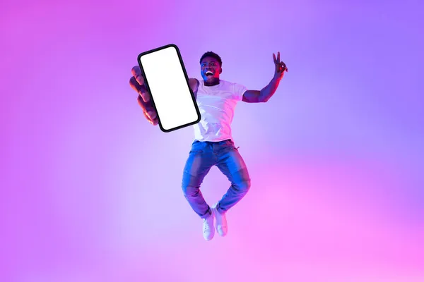 스마트폰으로 점프하는 흥분한 아프리카 계 미국인 남자의 전체 길이, 네온 라이트에 빈 화면, 흉내 — 스톡 사진