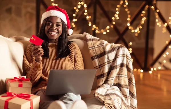 Julshopping. Svart kvinna med kreditkort och bärbar dator sitter på soffan hemma, köpa julklappar på webben hemma — Stockfoto