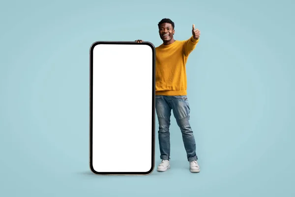 Положительный черный парень позирует с большим смартфоном, показывая большой палец вверх — стоковое фото