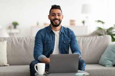 Bin yıllık neşeli Arap adam kanepesinde dizüstü bilgisayarla oturuyor, internet üzerinden çalışıyor ya da çalışıyor.