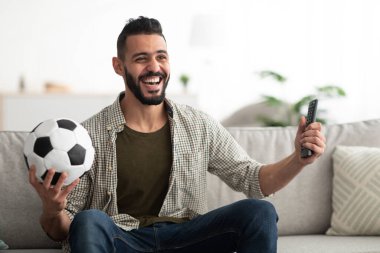 Televizyonda futbol maçı izleyen mutlu genç bir Arap adam, elinde top ve evinde kontrolör tutuyor.