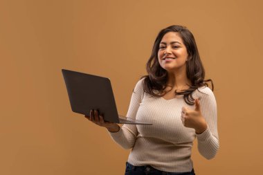 Öğrenciler için uzak işler. Mutlu Arap kadın dizüstü bilgisayar tutuyor ve baş parmağını kaldırıyor, kahverengi stüdyo geçmişi var.