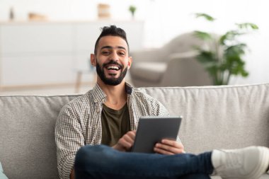Mutlu genç Arap adam dijital tablet kullanıyor, öğreniyor ya da uzaktan çalışıyor, evde online toplantı yapıyor.