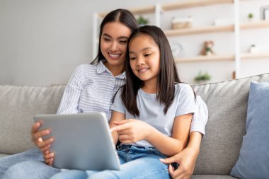 Kanepede oturup internette sörf yaparken dijital tablet kullanan heyecanlı Asyalı anne ve kız.
