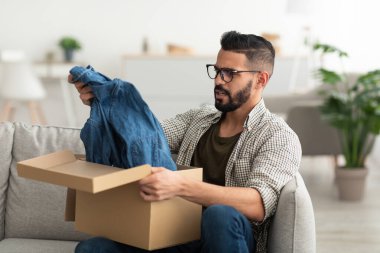 İnternetten alışveriş hatası. Mutsuz Arap adam karton paketi açıyor, kıyafet alıyor, teslimattan memnun değil.