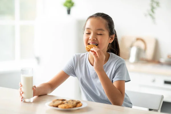 예쁘게 생긴 동양 여자가 과자를 먹고 부엌에서 우유를 마시고 행복 한 여자 아이는 집에서 건강에 좋은 간식을 즐긴다 — 스톡 사진