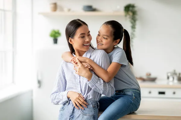 Concepto de maternidad. Bonita chica asiática abrazando a su joven madre desde la espalda, sentado en la cocina, espacio libre — Foto de Stock