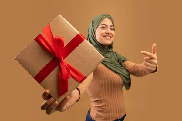 Positive arab lady holding wrapped present box and pointing on it, standing over brown background, selective focus — Stock Photo, Image