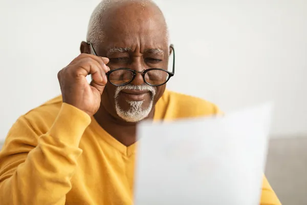 Hombre africano mayor entrecerrando los ojos leyendo papeles usando anteojos en interiores — Foto de Stock