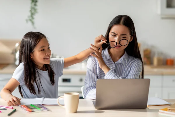 Азиатка работает за ноутбуком из дома, пока дочь отвлекает ее, снимая мамины очки — стоковое фото