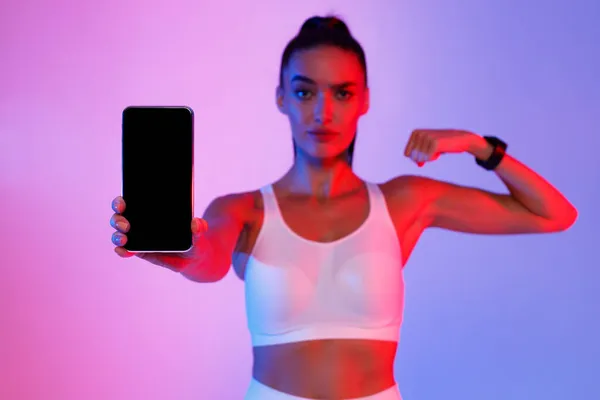 Athletic Woman Wyświetlanie ekranu smartfona i bicepsów na tle neonowym — Zdjęcie stockowe