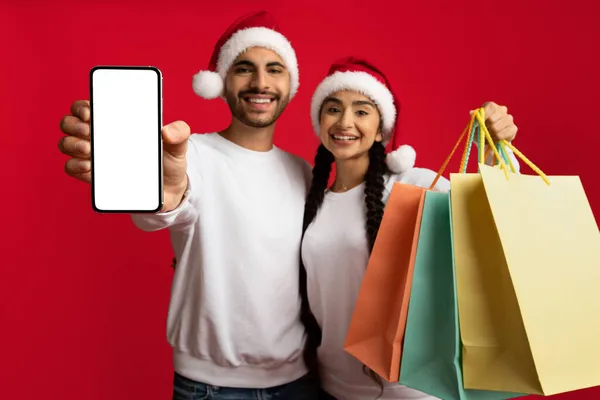 Noel Baba Şapkalı Orta Doğulu Çift Boş Akıllı Telefon ve Alışveriş Torbası Tutuyor — Stok fotoğraf