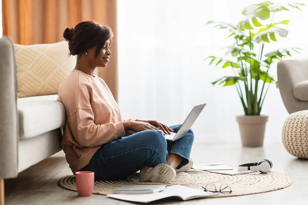 Ευτυχισμένη μαύρη γυναίκα που χρησιμοποιεί το φορητό υπολογιστή που εργάζονται σε απευθείας σύνδεση στο σπίτι, Side-View — Φωτογραφία Αρχείου