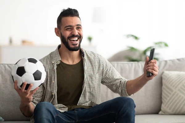 Ευτυχισμένος νεαρός Άραβας βλέποντας ποδόσφαιρο στην τηλεόραση, κρατώντας μπάλα και ελεγκτή στο σπίτι — Φωτογραφία Αρχείου