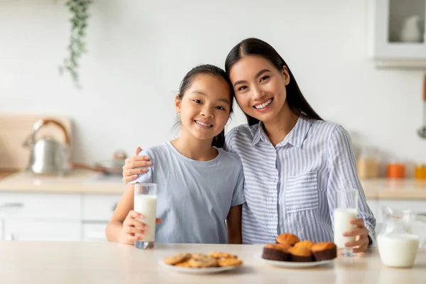 Genç Asyalı anne ve kız taze ev yapımı kurabiyelerin tadını çıkarıyor, mutfakta oturmuş süt içiyor. — Stok fotoğraf