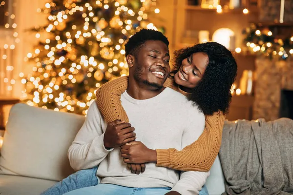 Affectueux jeune couple noir étreignant sur canapé dans une chambre décorée avec des lumières magiques à Noël — Photo