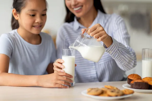 Ασιάτισσα μητέρα ρίχνει φρέσκο γάλα για τη χαριτωμένη έφηβη κόρη της, όμορφη κοπέλα παίρνει πρωινό με τη μαμά, κάθεται στην κουζίνα — Φωτογραφία Αρχείου