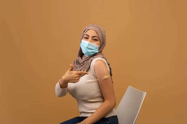 Senhora muçulmana vacinada feliz em hijab apontando para gesso no braço após a vacinação covid-19 bem sucedida, fundo marrom — Fotografia de Stock