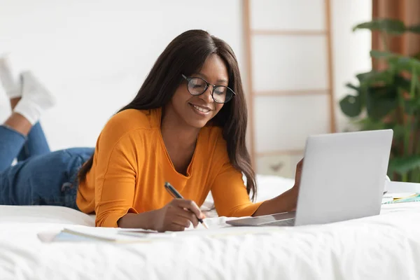 Nero donna apprendimento prendere note sdraiato con computer portatile in camera da letto — Foto Stock