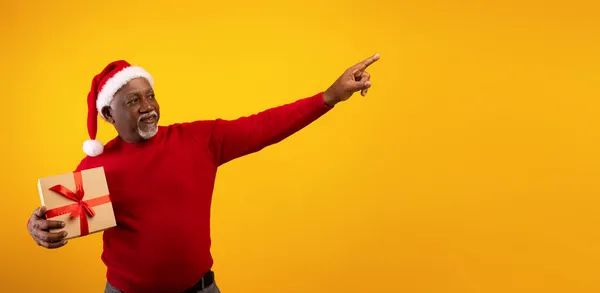 Feliz Navidad. Senior black man holding Xmas gift, pointing aside at empty space for your ad on orange background — Foto de Stock