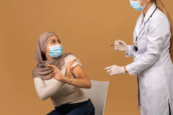 Mujer árabe joven con máscara facial asustada de hacer la inyección de la vacuna contra el coronavirus, haciendo un gesto STOP y mirando al médico —  Fotos de Stock