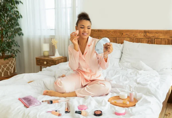 Dagelijkse schoonheidsroutine. Zwarte dame aanbrengen cosmetische foundation met applicator, kijken naar spiegel, zitten op bed — Stockfoto