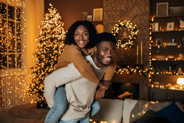 크리스마스 트리로 장식된 방에서 즐겁게 웃으며 여자 친구에게 피기백을 주는 행복 한 흑인 남자 — 스톡 사진