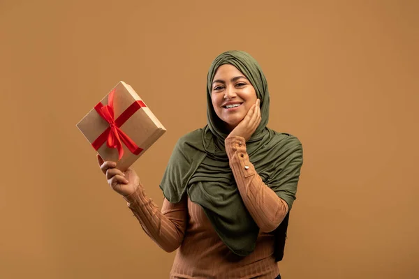 Happy muslim lady in hijab holding wrapped box, feeling excited on brown background, receiving birthday surprise — Stock Photo, Image