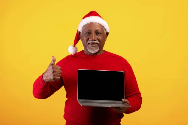 Sonriente anciano negro en Santa sombrero sosteniendo ordenador portátil con pantalla vacía y mostrando el pulgar hacia arriba gesto, maqueta — Foto de Stock