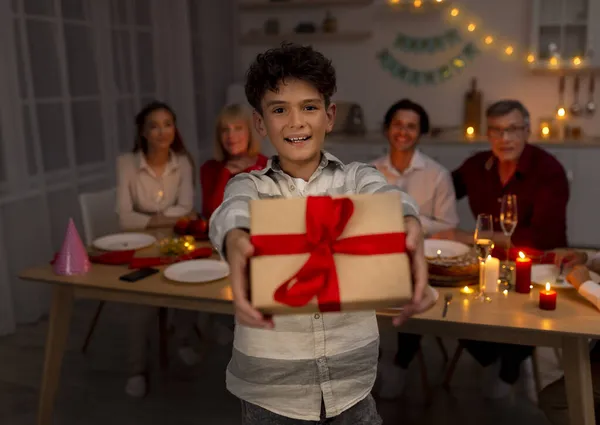 Happy little boy giving gift box at camera, having festive dinner at home — Stock Photo, Image