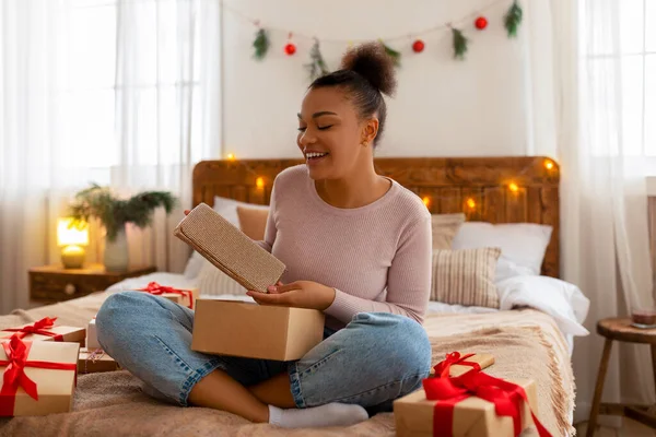 Sorpresa donna afroamericana disimballaggio scatole regalo di Natale e tenere la frizione, seduto sul letto e sorridente — Foto Stock