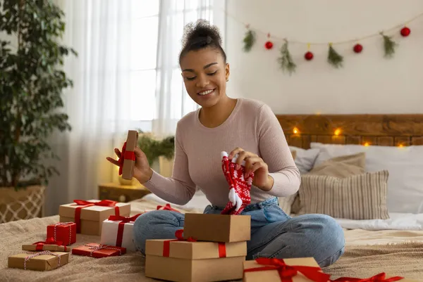 Positiva dama afroamericana tomando calcetines de la caja de regalo de Navidad, sentado en la cama en el dormitorio y desempacar regalos — Foto de Stock