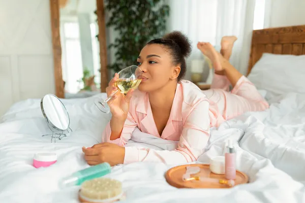 Senhora preta positiva em pijama beber vinho enquanto deitado na cama com produtos de cuidados de beleza ao lado dela — Fotografia de Stock