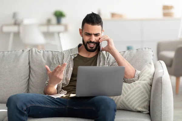 Schöner junger arabischer Mann telefoniert mit Smartphone, nutzt Laptop für Online-Treffen, arbeitet von zu Hause aus — Stockfoto