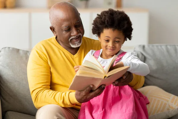 Alegre bonito afro-americano pequena neta e avô sênior leitura livro com contos de fadas no sofá — Fotografia de Stock