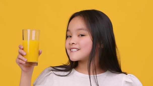 Přirozené vitamíny pro děti. Roztomilé malé asijské dívka drží sklenici čerstvé pomerančové šťávy a kývnutí na kameru — Stock video