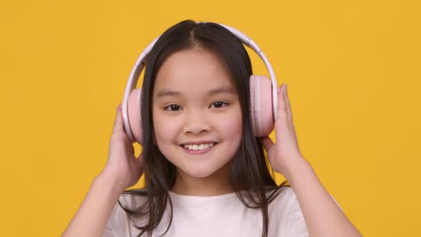 Portré aranyos kis ázsiai lány visel nagy vezeték nélküli fejhallgató mosolyog és kacsint a kamera, élvezi a zenei szerkentyű — Stock videók