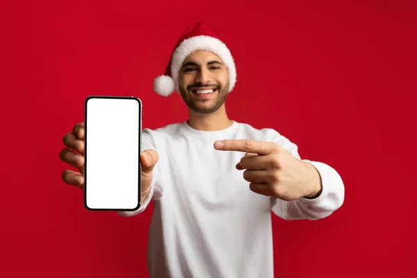 Çevrimiçi teklif. Noel şapkalı neşeli Arap adam boş akıllı telefonu işaret ediyor. — Stok fotoğraf