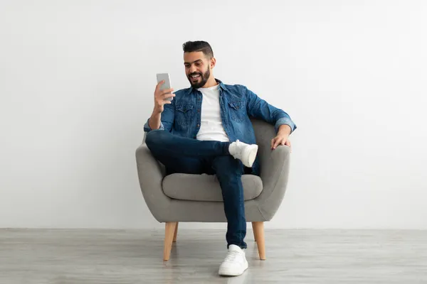 Glücklicher arabischer Millennial-Mann sitzt im Sessel mit Smartphone, schreibt SMS an Freund, verschickt SMS, surft im Web — Stockfoto
