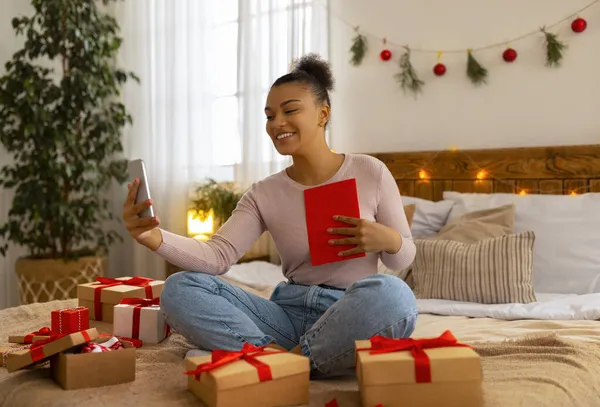 Happy african american lady holding greeting card and talking via smartphone video call, sitting on bed in cozy bedroom
