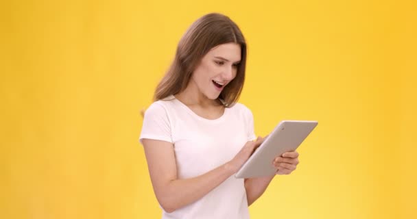 Olumlu haber. Dijital tablette sosyal medya okuyan ve gülümseyen, sarı stüdyo geçmişi olan neşeli genç bir kadın. — Stok video