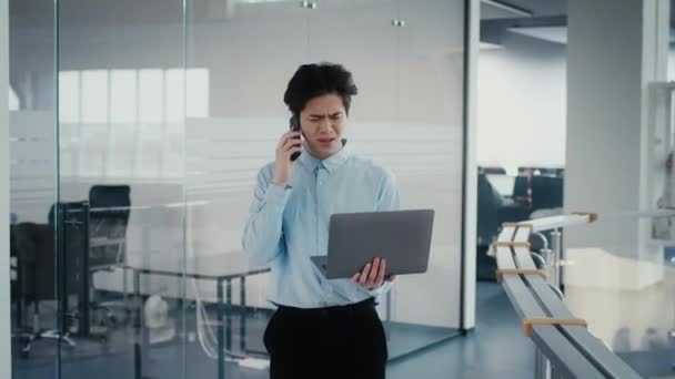 Uomo d'affari asiatico scontento che utilizza il computer portatile che parla al telefono in ufficio — Video Stock