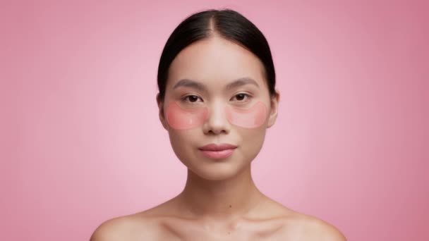 Mujer asiática con parches aplicados debajo de los ojos Cara hidratante, fondo rosa — Vídeos de Stock