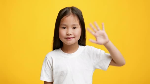 Маленькая корейская девочка машет рукой, показывая "Hello Over Yellow Background" — стоковое видео