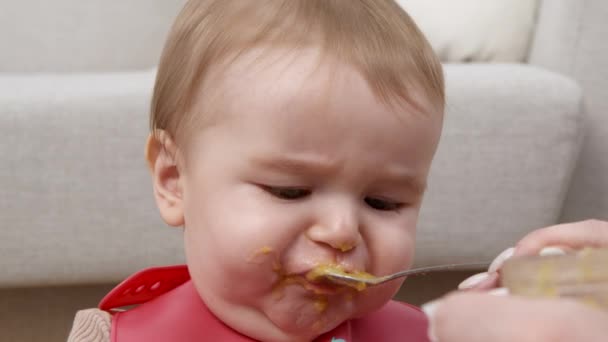 Első kisgyermek táplálkozás. Imádnivaló kisbaba partedlit visel, organikus pürét eszik, anya kanállal eteti a gyereket. — Stock videók
