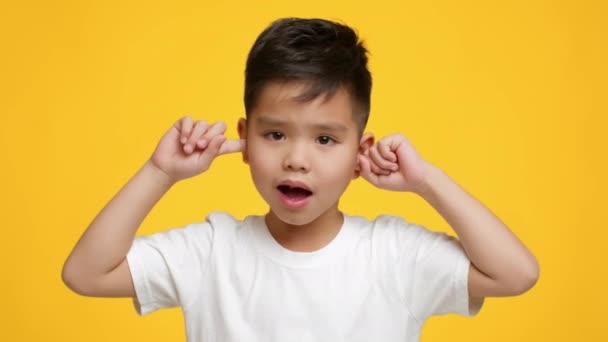 Travieso asiático pequeño niño cubriendo orejas sacudiendo cabeza, fondo amarillo — Vídeo de stock