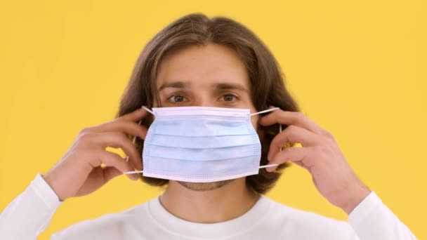 Personal preventive measures. Young man putting on protective medical mask, looking at camera over yellow background — Stock Video