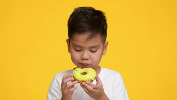 Little Asian Boy Eating Tasty Donut Standing Over Yellow Background — Stock Video